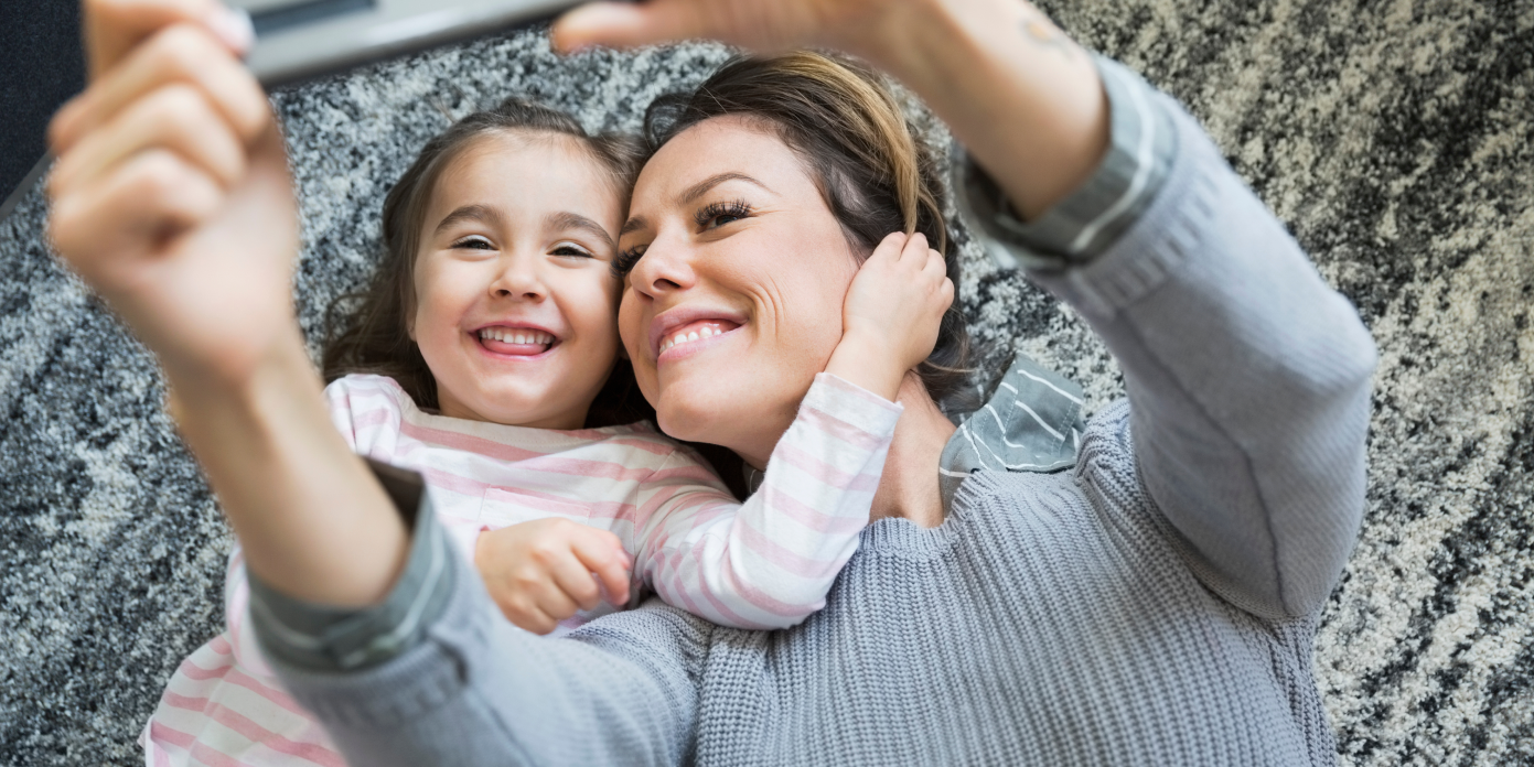 Woman and girl laying down, looking at a phone and smiling